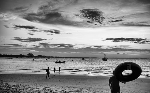 Black and white photography of tourist bathers at a brazilian northeastern beach. A man in swimsuit carries a tire float towards the sea