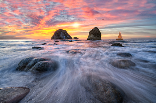 Water Is Flowing Over Coastal Sea Rocks With A Sailboat And Sunset Sky High Resolution