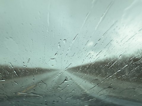 scene from inside the car looking at the road ahead with the rain splatting on the windshield. Sky is gray with clouds and the road is out of focus