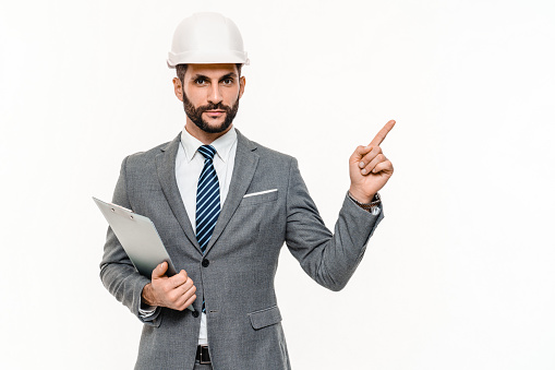 Successful young engineer in white helmet and clipboard pointing on copyspace isolated over white. Foreman construction worker real estate agent showing background for blank space