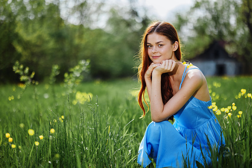 Woman in the springtime nature smile with teeth sitting in a field on the green grass with a yellow flower in her hand and smiling happy sunset in a blue dress with red hair, protection from insects. High quality photo