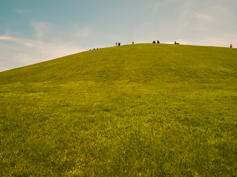 Tall green hill covered in lush green grass against a cloudy blue sky. Silhouettes of unrecognizable people gathered at the top of the hill for a viewing party.