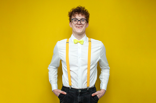 young guy in festive outfit and glasses, nerd student in bow-tie shirt and suspenders stands on yellow isolated background