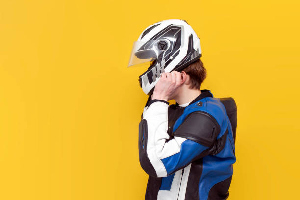 young guy motorcyclist in leather jacket puts on helmet on yellow isolated background, professional racer in protective gear holds helmet, copy space