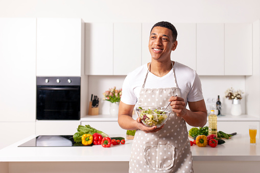 young african american man in apron holds plate of veggie salad in white modern kitchen and looks away at the copy space, home cook guy advertises healthy meal of vegetables and herbs