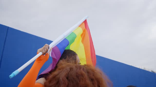 Young smiling drag queen waving rainbow flag on gay pride festival day in front of a blue wall
