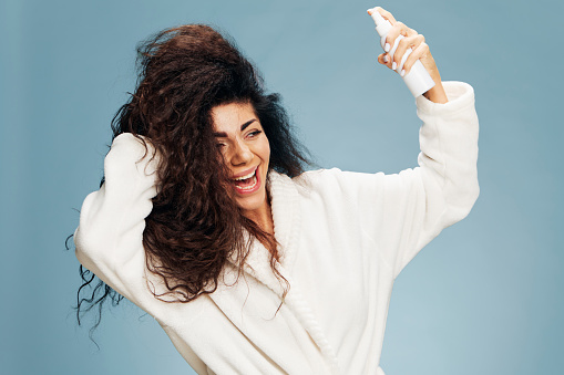 Hairstyling morning routine. Overjoyed tanned curly Latin lady in bathrobe Spraying On Hair For Repair split ends posing isolated on pastel blue background, smiling aside. Copy space offer