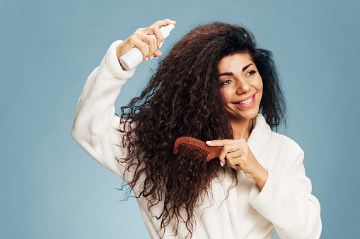 Hairstyling routine. Overjoyed tanned curly Latin lady in bathrobe Spraying On Hair For Repair split ends posing isolated on pastel blue background, using hairbrush, smiling. Copy space offer