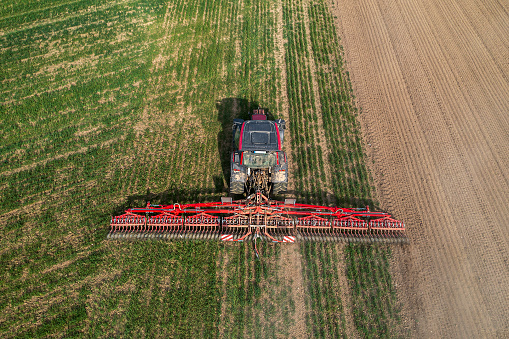 Aerial view of a red modern agricultural tractor cultivating field with a large rotary harrow in spring.