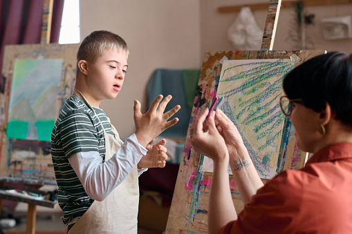 Portrait of young boy with Down syndrome choosing paint colors while painting on easel in art class with teacher helping