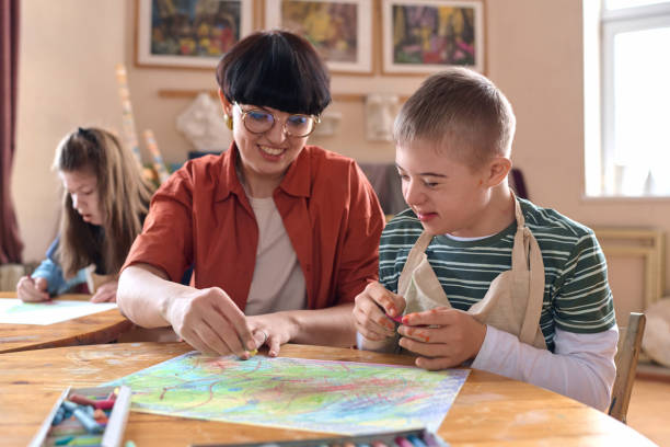 Smiling Boy with Disability Enjoying Art Class in Studio with Mentor Assisting Portrait of smiling boy with disability enjoying art class in studio with mentor assisting community outreach stock pictures, royalty-free photos & images