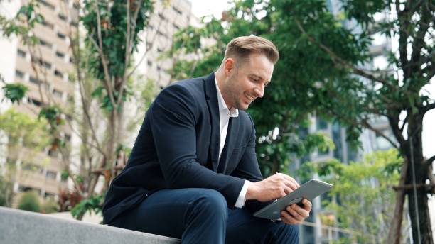 istock Businessman sitting at park while working on tablet by reading report. Urbane 2148218424