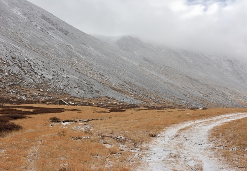 A field road covered with snow turns right along a high mountain range on a cloudy autumn morning.