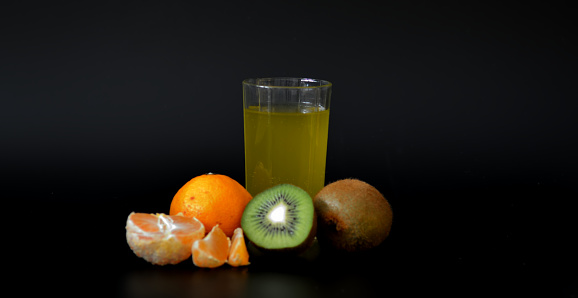 A glass of a mixture of tropical fruits with seeds on a black background, next to a ripe tangerine and kiwi. Close-up.