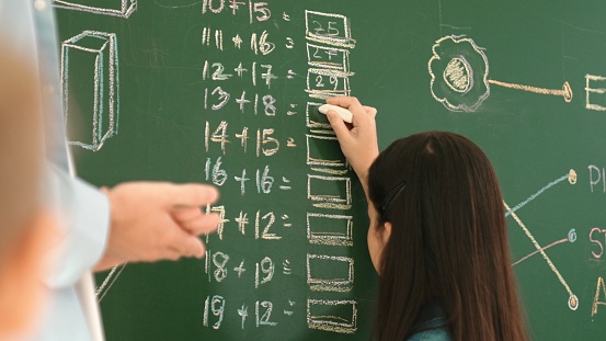 Young asian girl writing math formula at blackboard while standing at classroom. Attractive elemental student studying or learning at math lesson while answer question. Education concept. Pedagogy.