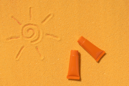 Small orange tubes of sunscreen and the sun painted on the sand. A preparation to protect the skin from sunlight.