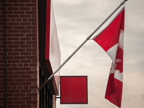 This image presents a compelling visual of an empty red signboard ready for customization, set against the textured backdrop of a brick wall facade. Above it, the iconic Canadian flag waves proudly, emblematic of the country's rich heritage and the unity of its people. The red of the sign complements the flag, hinting at the strong sense of identity and pride that characterizes Canadian businesses. This scene, capturing the essence of retail and commerce in Canada, offers ample space for business owners to display their brand, inviting passersby to envisage the potential within.