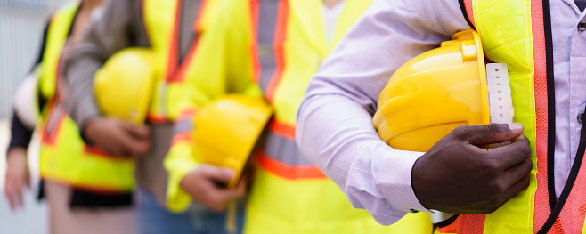 Close up shot of engineer or foreman team holding a protective safety helmet beside their body. A shipyard - container yard workers and logistic engineers holding a yellow safety hardhat or helmet.