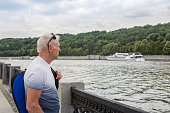 A sporty gray-haired man with a blue sports bag, wearing a T-shirt, stands on the river embankment on a cloudy day