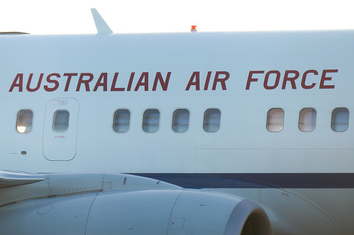 The central fuselage and engine of a Royal Australian Air Force Boeing B737-7DT (BBJ) plane, registration A36-001, used for VIP transport such as the Governor-General and Prime Minister.   She is parked at Sydney Kingsford-Smith Airport. This image faces west and the setting sun and was taken from Ross Smith Avenue, Mascot, behind a steel security fence, on a hot and sunny afternoon on 19 January 2024.