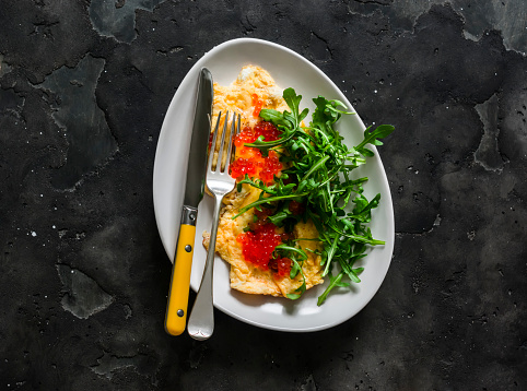 Cheese omelet with red caviar and arugula on a dark background, top view