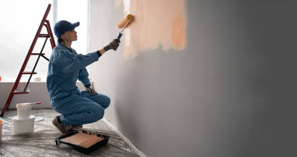 female painter in overalls with cap and gloves painting a wall with paint roller, ladder and buckets on background. Copy space female painter in overalls with cap and gloves painting a wall with paint roller, ladder and buckets on background. Side view. Copy space house painting service stock pictures, royalty-free photos & images