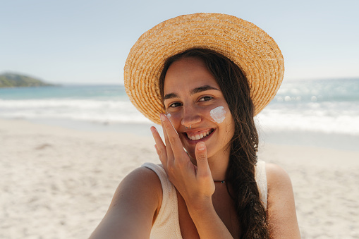 Photo of a young woman applying sunscreen on her face while at the beach