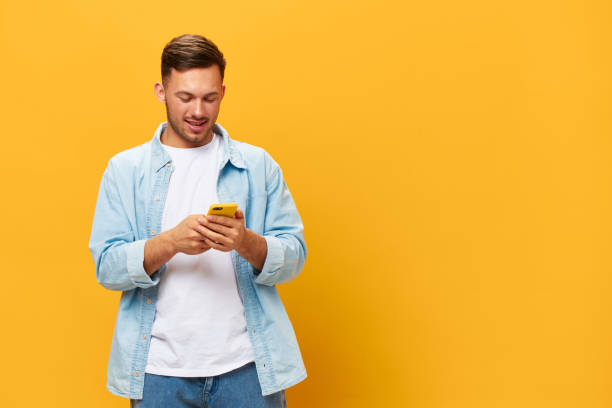 istock Happy smiling tanned handsome man in blue basic t-shirt type message chats with friends using phone posing isolated on yellow studio background. Copy space Banner Mockup. Online Social media concept 2148230909