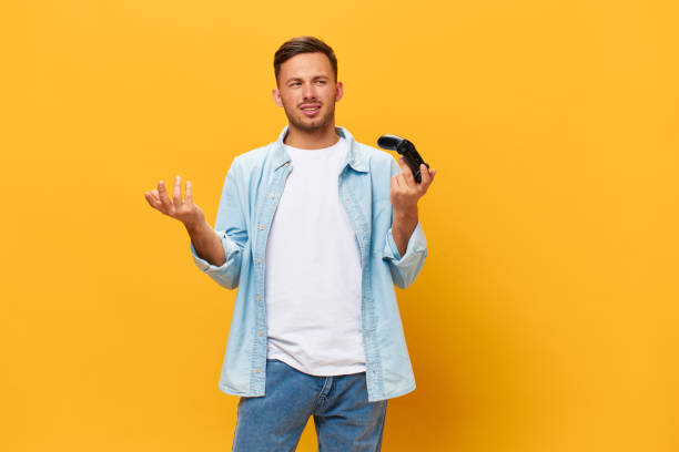 istock Confused unhappy tanned handsome man in blue basic t-shirt raise hands up with joystick gamepad posing isolated on orange yellow studio background. Copy space Banner Mockup. People lifestyle concept 2148231124