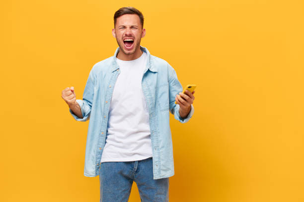istock Angry irritated tanned handsome man in blue basic t-shirt scream at camera hold phone posing isolated on orange yellow studio background. Copy space Banner Mockup. Online Social media concept 2148231164