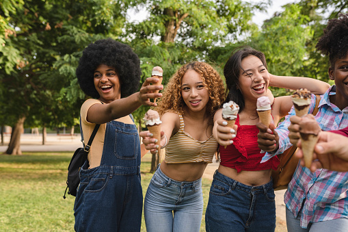 Group of joyful friends sharing a playful moment, extending their ice cream cones towards the camera in a park.
