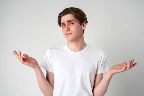 istock Young Man in White T-Shirt Displaying Uncertain Gesture Against Light Background 2148233378