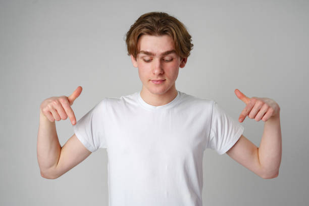 Young Man in White T-Shirt Pointing Downwards With Both Hands Against Plain Background