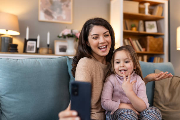 istock Smiling mother and daughter on video call using digital phone at home. while making video call and waving hello sitting on sofa. Woman and her cute little kid doing video conference with grandparents. 2148234207