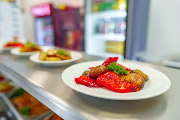 A variety of carefully prepared gourmet dishes are presented on white plates, placed on the shelves of a deli. The focus is on a plate of charred vegetables at the forefront, with other savory items in the background, suggesting a selection of ready-to-eat meals.