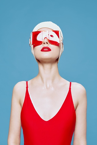 A bright red-lipped lady in a red swimsuit glasses swimming cap raises her head up posing isolated on a blue background in the studio. High quality photo