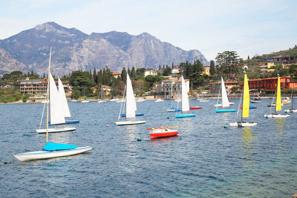 A fleet of sailboats engaging in sport on the sparkling waters of Lake Garda, with the lush landscape of the Italian countryside and mountains in the background