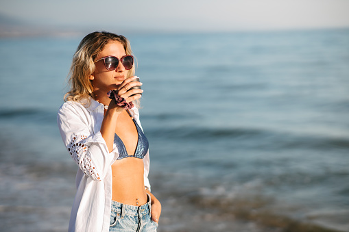 Morning coffee by the beach. Beautiful girl in white shirt drinking coffee by the beach