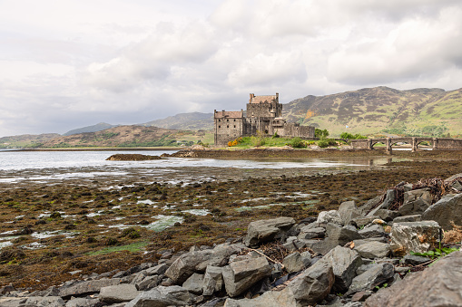 Dornie - United Kingdom. May 22, 2023: Eilean Donan Castle, overlooking a seaweed-strewn loch, is framed by a rugged highland backdrop and an arching stone bridge, encapsulating Scotland timeless allure