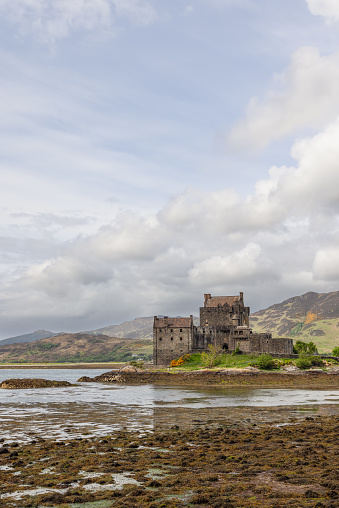 Dornie - United Kingdom. May 22, 2023: Eilean Donan Castle, poised on the edge of a loch, showcases the rugged beauty and heritage of the Highlands, with a serene sky and low tide revealing the algae-covered shore (Vertical photo)