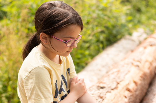 A little cute girl with glasses and a ponytail sits sadly on sawn tree trunks