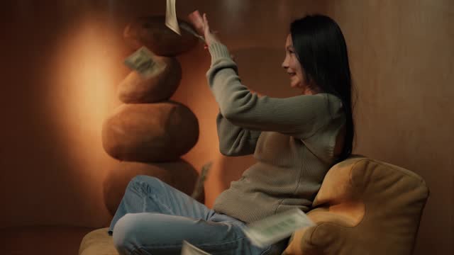 Asian Woman Joyfully Throwing Cash in the Air Sitting on a Couch Indoors
