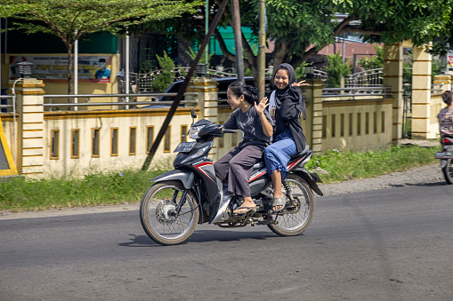 Kuala, North Sumatra, Indonesia - January 25th 2024: Two young women having fun on a motorcycle in the street in a small Indonesian village