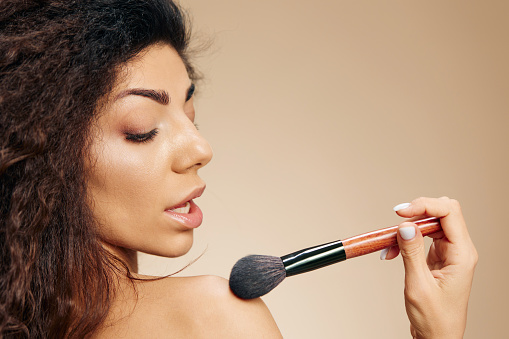 VISAGE CONCEPT. Profile sexy adorable tanned awesome curly Latin lady hold makeup brush on shoulder apply powder highlighter posing isolated on pastel beige background look down. Copy space banner