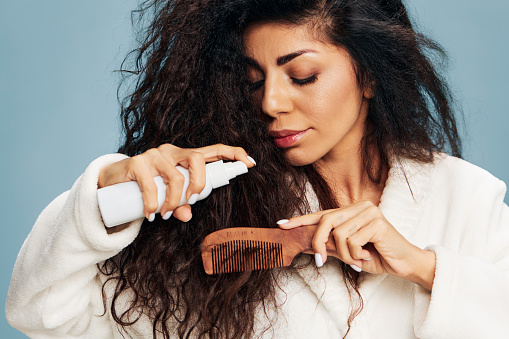 Haircare And Hairstyling. Smiling Pensive tanned curly Latin lady in bathrobe Spraying On Hair For Repair split ends posing isolated on pastel blue background, using hairbrush. Hair Care Ad Concept