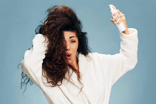 Hairstyling morning routine. Excited shocked tanned curly Latin lady in bathrobe Spraying On Hair For Repair split ends posing isolated on pastel blue background, touching hair. Copy space offer