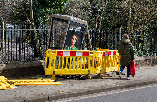 A woman passing a damaged telephone box, surrounded by yellow safety barriers, on the pavement in a street in Penge, South East London. It has been struck by a vehicle travelling at some speed.