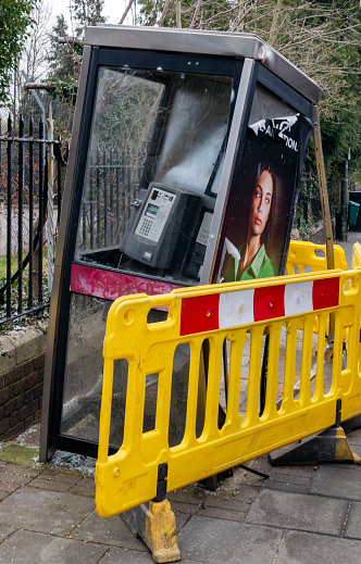 A damaged telephone box, surrounded by yellow safety barriers, on the pavement in a street in Penge, South East London. It has been struck by a vehicle travelling at some speed.