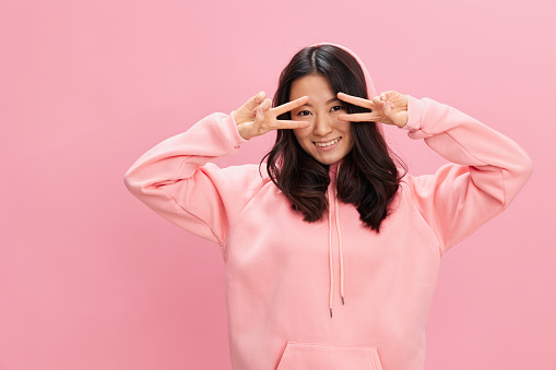 Lovely Asian student young lady in pink hoodie sweatshirt show v-sign gesture smile at camera posing isolated on over pink studio background. Good offer. Fashion New Collection concept