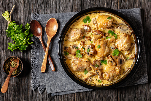chicken thighs in a creamy mushroom garlic sauce with herbs and parmesan cheese in baking dish on dark wooden table with wooden spoons and napkin, horizontal view from above, flat lay